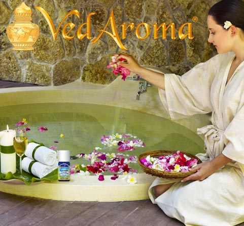 A woman in a bathrobe drops flower petals into a large sunken bath tub as she prepares to relax with VedAroma essential oils applied to her bath.